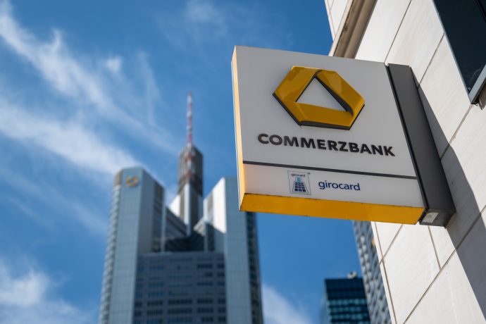 Commerzbank-Zeichen auf einem Gebäude vor dem Hintergrund eines modernen Wolkenkratzers und eines klaren blauen Himmels.