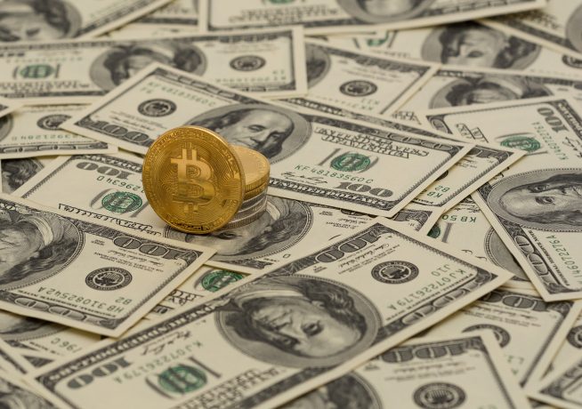 Eine goldene Bitcoin-Münze, die den Krypto-Reichtum symbolisiert, steht aufrecht auf einem verstreuten Stapel von Hundert-Dollar-Scheinen.
