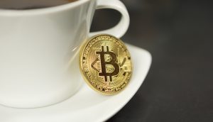 Ein goldfarbener Bitcoin-Token ruht auf einer weißen Kaffeetasse auf einer Untertasse, mit Kaffee in der Tasse und einer dunklen Oberfläche im Hintergrund - eine moderne Anspielung auf Krypto-Fans.
