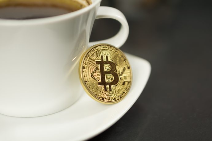 Ein goldfarbener Bitcoin-Token ruht auf einer weißen Kaffeetasse auf einer Untertasse, mit Kaffee in der Tasse und einer dunklen Oberfläche im Hintergrund - eine moderne Anspielung auf Krypto-Fans.