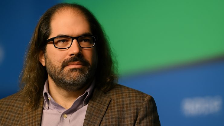 Ein Mann mit langen Haaren und Bart, der eine Brille, einen karierten Blazer und ein Kragenhemd trägt, sitzt vor einem blau-grünen Hintergrund mit dem XRP-Logo.