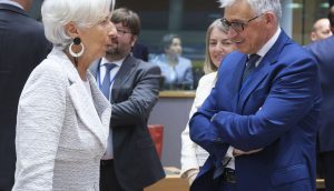 Zwei förmlich gekleidete Personen diskutieren in einem Konferenzraum über Euro-Strategien, während mehrere andere Personen im Hintergrund stehen und sich unterhalten.