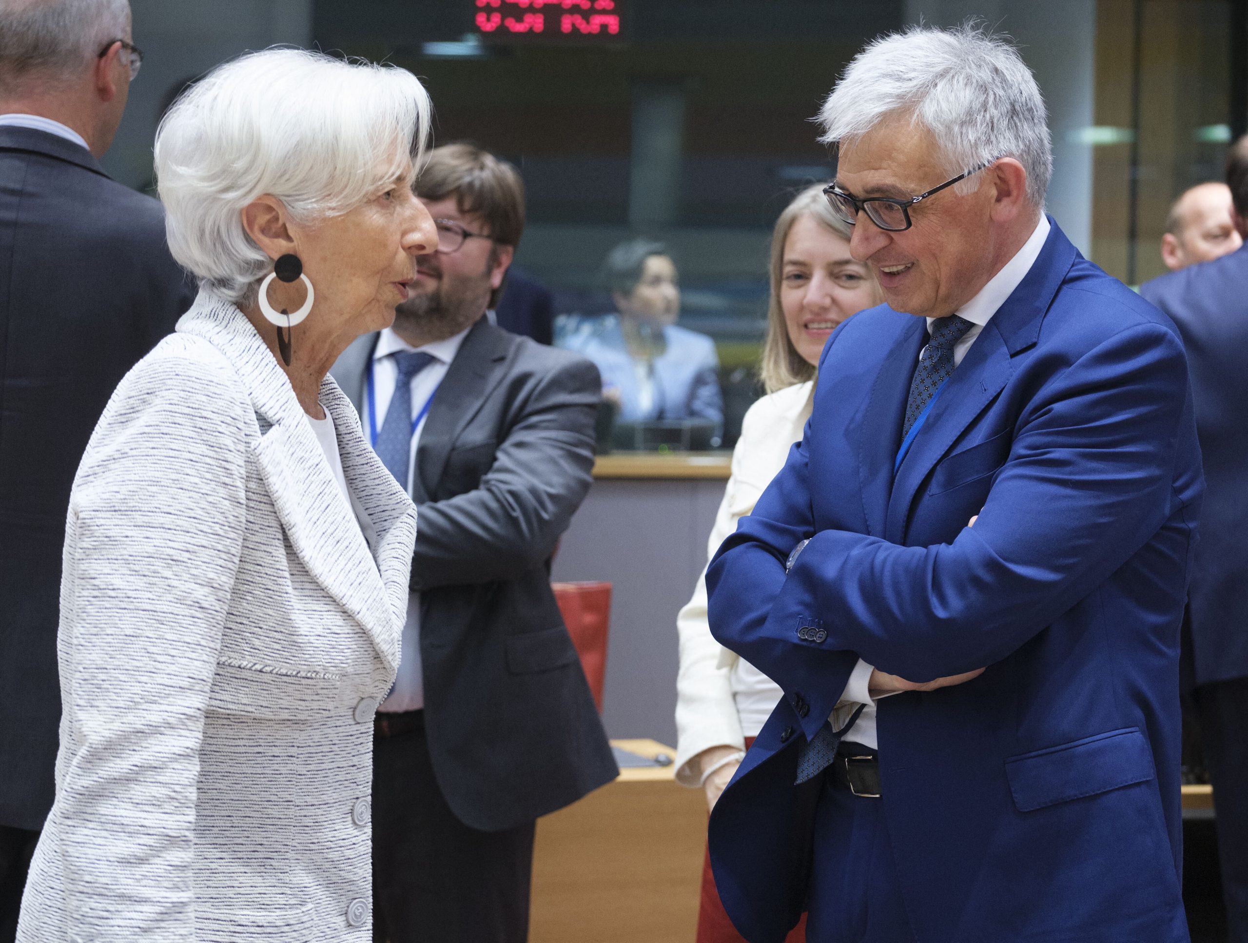 Zwei förmlich gekleidete Personen diskutieren in einem Konferenzraum über Euro-Strategien, während mehrere andere Personen im Hintergrund stehen und sich unterhalten.
