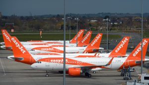 Mehrere EasyJet-Flugzeuge, eine Anspielung auf die Vision von EasyJet-Gründer, sind an einem klaren Tag an den Gates des Flughafens geparkt, mit Flughafeninfrastruktur und Landschaft im Hintergrund.