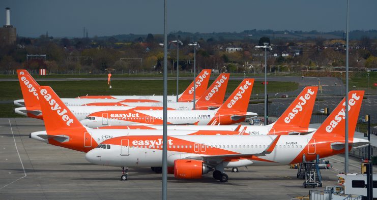 Mehrere EasyJet-Flugzeuge, eine Anspielung auf die Vision von EasyJet-Gründer, sind an einem klaren Tag an den Gates des Flughafens geparkt, mit Flughafeninfrastruktur und Landschaft im Hintergrund.