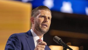 Eric Trump, ein Mann in einem blauen Anzug und einer rot gestreiften Krawatte, der Trump ähnelt, spricht auf einer Bühne in ein Mikrofon, vor einem verschwommenen Hintergrund mit blauen und gelben Lichtern.