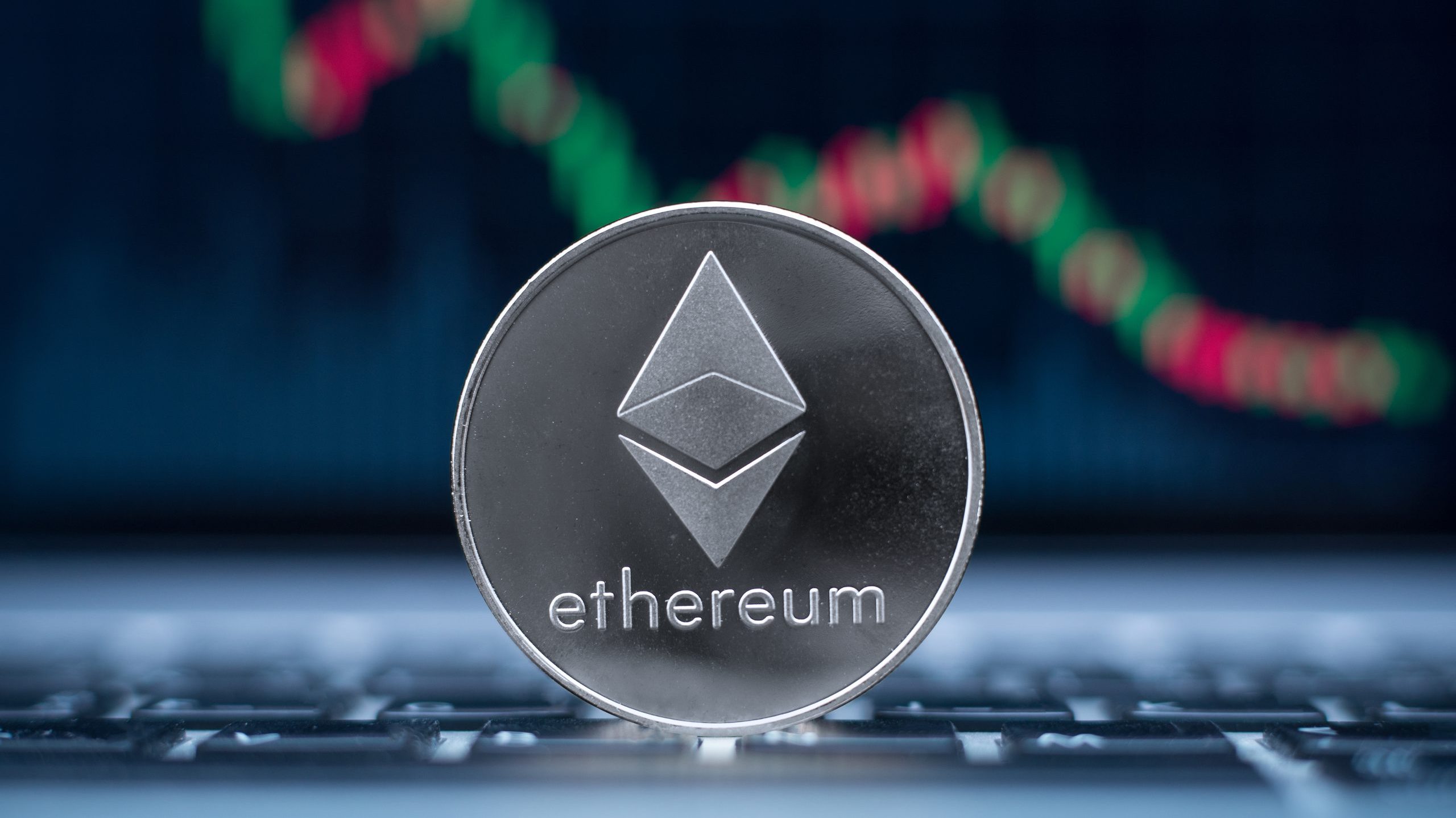 Eine silberne Ethereum-Münze steht auf einer Laptop-Tastatur mit einem verschwommenen roten und grünen Candlestick-Chart im Hintergrund und symbolisiert die Ethereum-Reserven an Börsen auf ihrem niedrigsten Stand.
