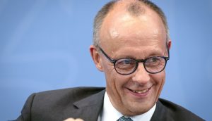 Ein Mann mittleren Alters im Anzug und mit Brille lächelt, während er vor einem blauen Hintergrund sitzt, der an den professionellen Geist der deutschen Finanzwelt und die moderne Welt des Bitcoin erinnert.