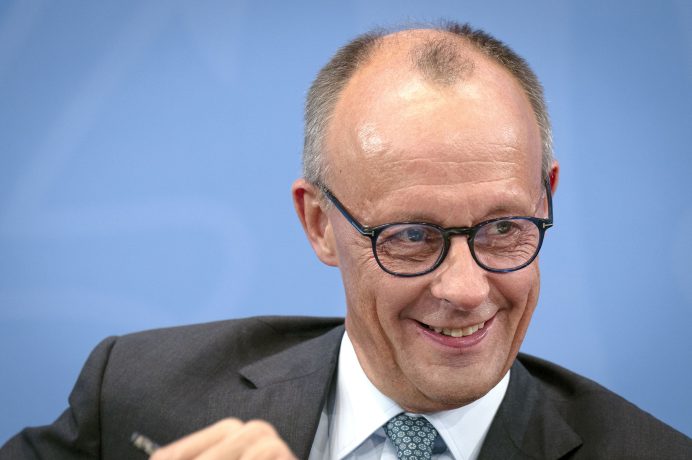 Ein Mann mittleren Alters im Anzug und mit Brille lächelt, während er vor einem blauen Hintergrund sitzt, der an den professionellen Geist der deutschen Finanzwelt und die moderne Welt des Bitcoin erinnert.