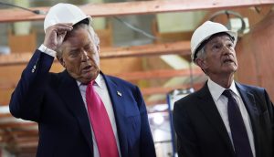 Jerome Powell und Donlad Trump in Anzügen und mit weißen Bauhelmen stehen in einem Haus; einer justiert seinen Helm, während der andere nach oben blickt und den Fortschritt eines Fed-Projekts zu beurteilen scheint.