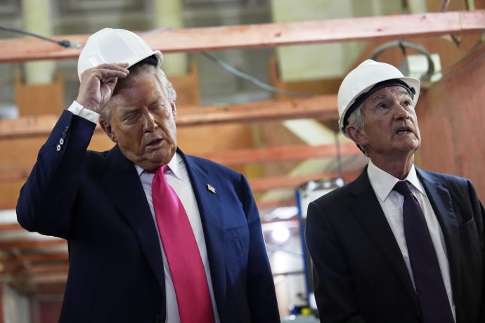 Jerome Powell und Donlad Trump in Anzügen und mit weißen Bauhelmen stehen in einem Haus; einer justiert seinen Helm, während der andere nach oben blickt und den Fortschritt eines Fed-Projekts zu beurteilen scheint.