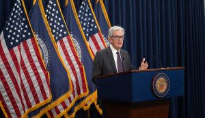 Ein Mann im Anzug spricht auf einem Podium mit dem Siegel der Federal Reserve vor vier US-Flaggen und blauen Vorhängen. Er äußert Bedenken über eine mögliche Zinssenkung und warnt davor, einen historischen Fehler zu machen.