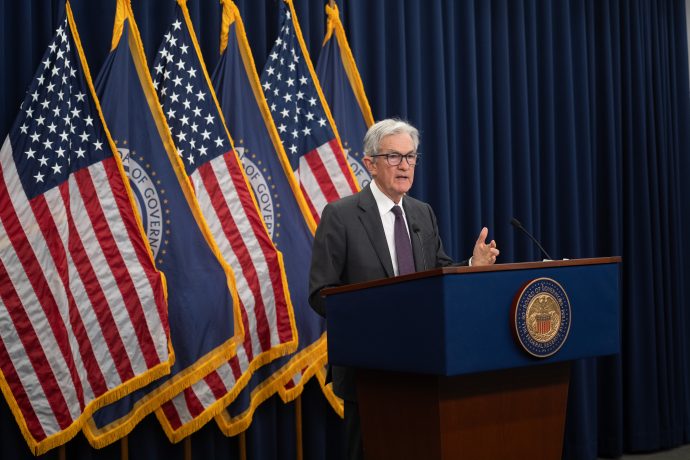 Ein Mann im Anzug spricht auf einem Podium mit dem Siegel der Federal Reserve vor vier US-Flaggen und blauen Vorhängen. Er äußert Bedenken über eine mögliche Zinssenkung und warnt davor, einen historischen Fehler zu machen.