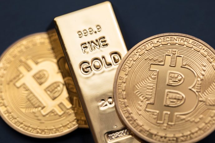 Nahaufnahme eines Goldbarrens und zweier physischer Bitcoin-Münzen, die nebeneinander auf einem dunklen Hintergrund platziert sind, um den Kontrast zwischen Gold und digitaler Währung hervorzuheben.