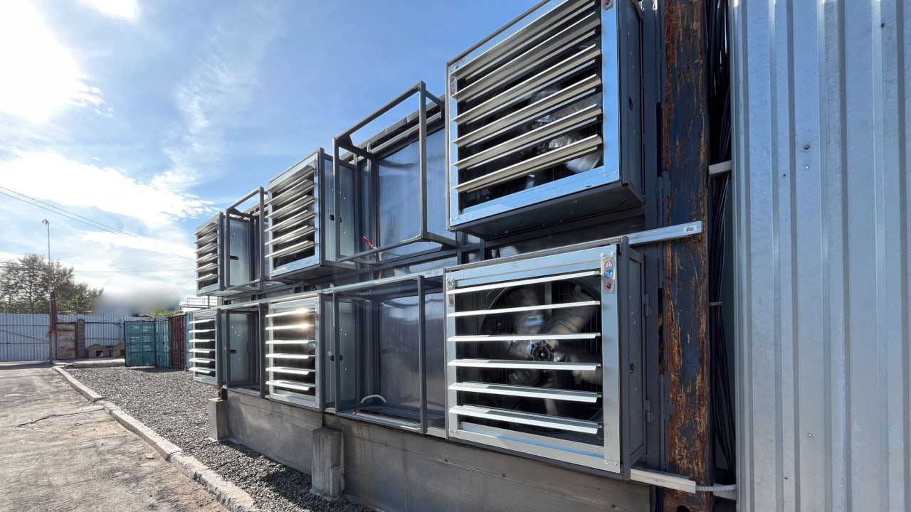 image An der Außenwand eines Metallgebäudes sind mehrere große Test-Power-Industrieventilatoren angebracht, die auf mögliche Bitcoin-Mining-Aktivitäten hindeuten, während in der Nähe ein Schotterplatz und Frachtcontainer zu sehen sind.