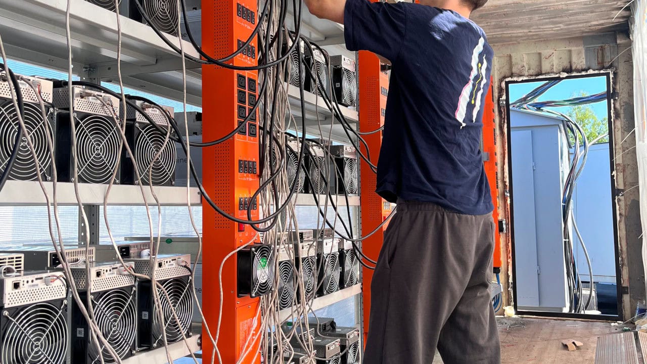 image Eine Person schließt Kabel an orangefarbene Racks mit mehreren Bitcoin-Mining-Maschinen in einem Raum mit freiliegenden Wänden und Kabeln an und bereitet sich auf das kostenlose Ausprobieren des Test-Power-Setups vor.