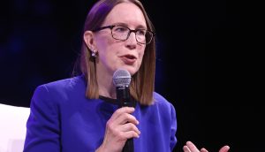 SEC-Kommissarin Hester Peirce leitet die Krypto-Taskforce Eine Frau mit glattem braunem Haar, Brille und blauem Blazer spricht in ein Mikrofon über Bitcoin, während sie vor einem dunklen Hintergrund sitzt.