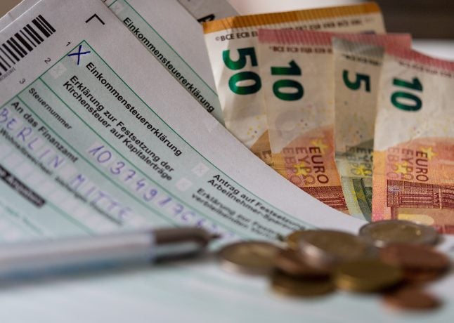 Deutsche Einkommensteuerformulare, Euro-Banknoten, ein Stift, Euro-Münzen und Krypto-Dokumente liegen auf einem Schreibtisch.