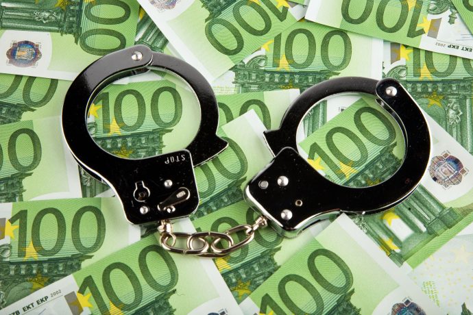 Ein Paar Metallhandschellen liegt offen auf mehreren 100-Euro-Banknoten und deutet auf die Überschneidung von Steuerhinterziehung und Krypto hin.