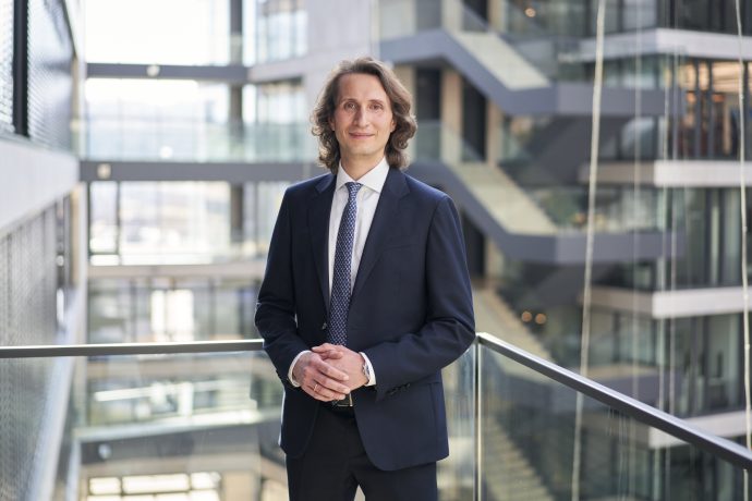 Ein Mann in einem dunklen Anzug steht in einem modernen Bürogebäude an einem Glasgeländer und denkt über steuereffiziente Lösungen für die Krypto-Erbschaft nach, inmitten der eleganten Treppenhäuser im Hintergrund.