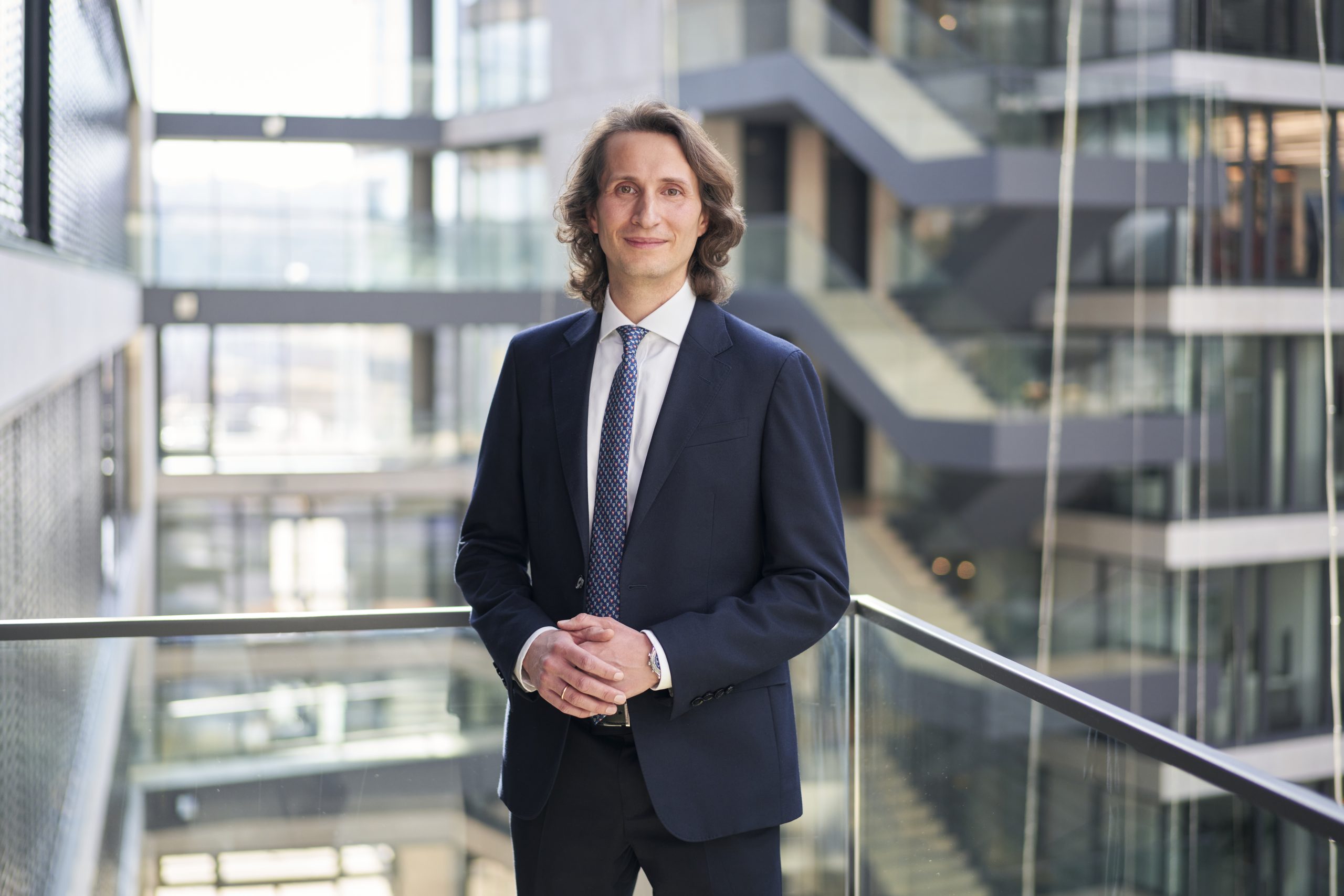 Ein Mann in einem dunklen Anzug steht in einem modernen Bürogebäude an einem Glasgeländer und denkt über steuereffiziente Lösungen für die Krypto-Erbschaft nach, inmitten der eleganten Treppenhäuser im Hintergrund.
