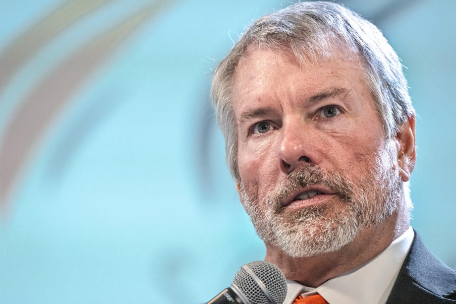 Ein Mann mittleren Alters mit grauem Haar und Bart spricht in einem Anzug und mit orangefarbener Krawatte vor einem unscharfen Hintergrund über Bitcoin in ein Mikrofon.