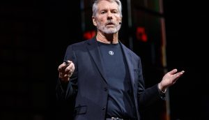Ein Mann mit grauem Haar und Bart, der einen schwarzen Anzug und ein T-Shirt trägt, spricht auf der Bühne über ein Headset-Mikrofon über Strategie und gestikuliert mit seinen Händen.