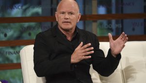 Ein Mann in einer schwarzen Jacke sitzt auf einer weißen Couch und gestikuliert mit seinen Händen, während er in einem Interview über Bitcoin spricht.