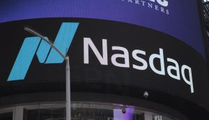 Ein markantes Nasdaq-Logo und Text leuchten auf einem elektronischen Bildschirm in einer belebten städtischen Umgebung, vor der eine Straßenlaterne steht.