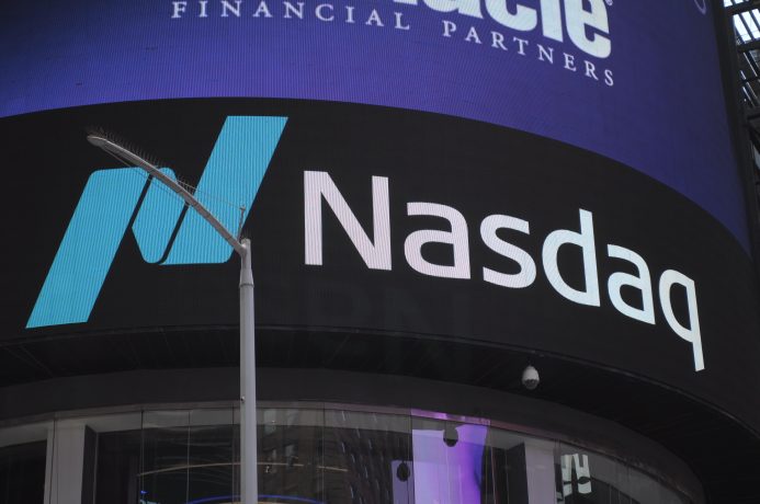 Ein markantes Nasdaq-Logo und Text leuchten auf einem elektronischen Bildschirm in einer belebten städtischen Umgebung, vor der eine Straßenlaterne steht.