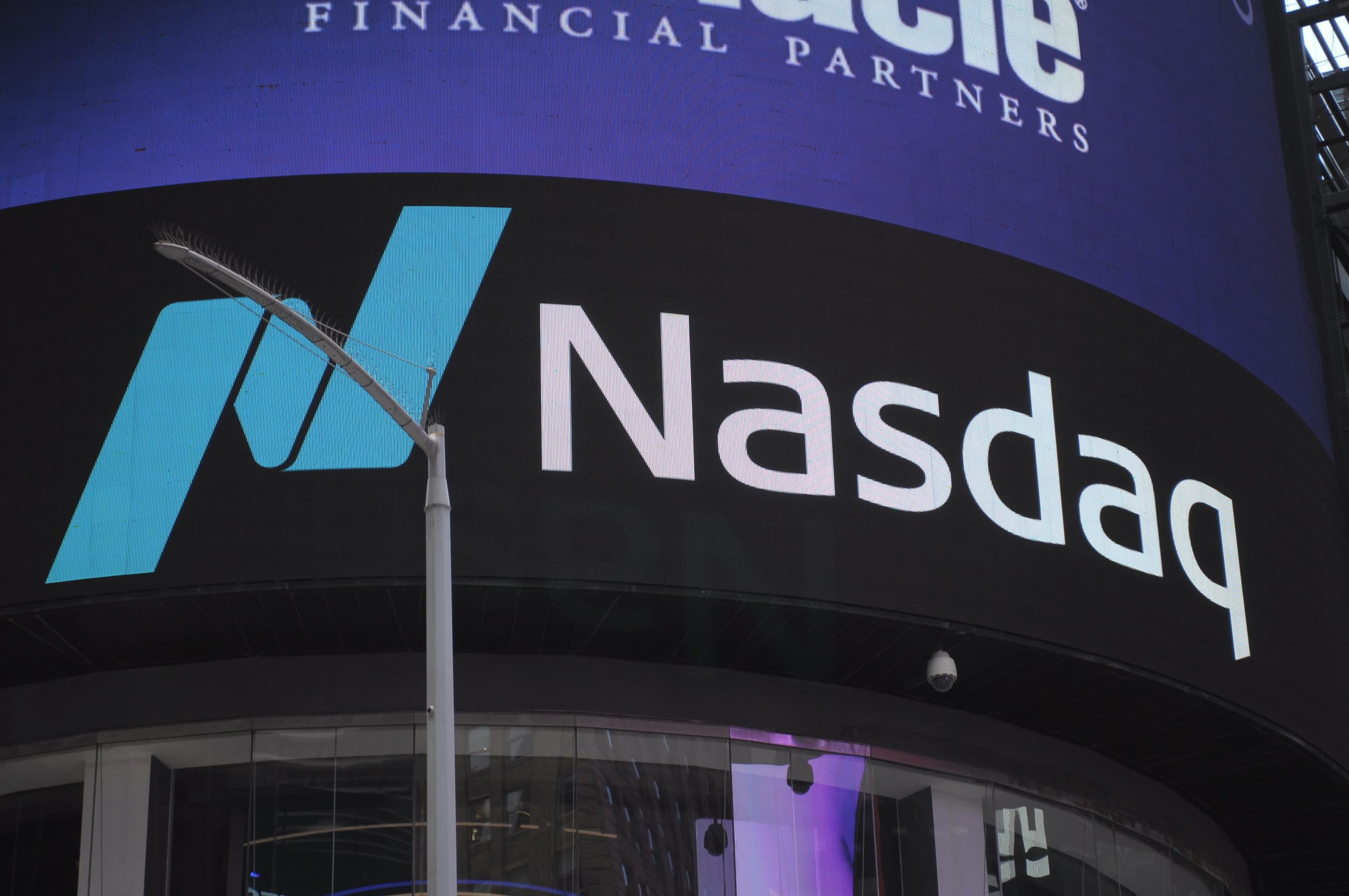 Ein markantes Nasdaq-Logo und Text leuchten auf einem elektronischen Bildschirm in einer belebten städtischen Umgebung, vor der eine Straßenlaterne steht.