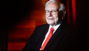 Warren Buffett, ein älterer Mann in Anzug und roter Krawatte, sitzt vor einem roten Hintergrund, blickt leicht zur Seite und lächelt.