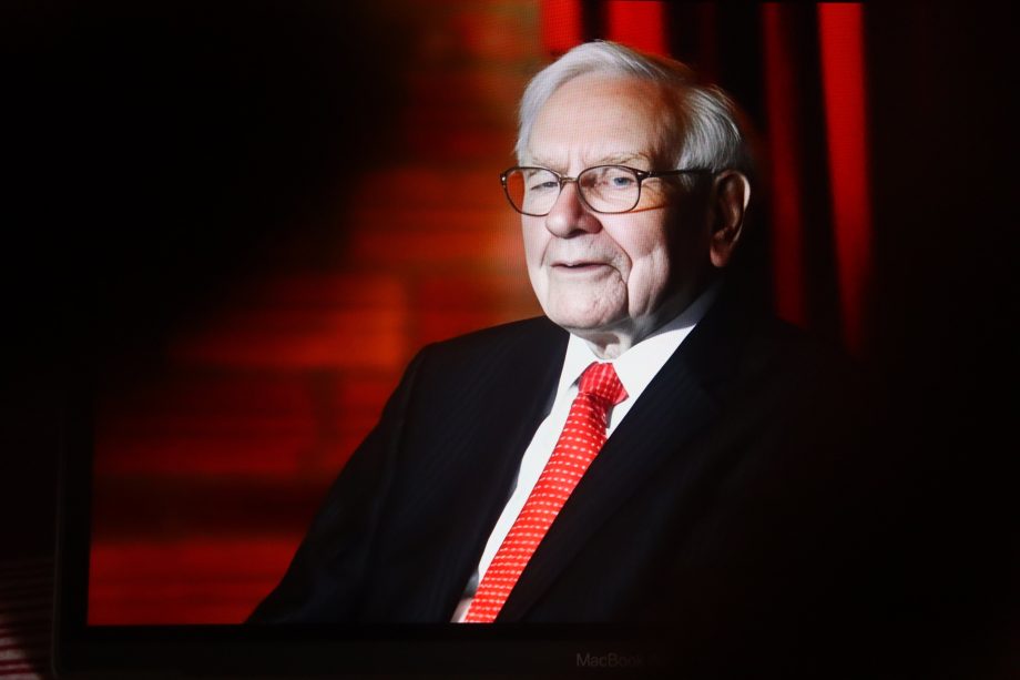 Warren Buffett, ein älterer Mann in Anzug und roter Krawatte, sitzt vor einem roten Hintergrund, blickt leicht zur Seite und lächelt.