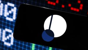 Ein Smartphone mit automatisch gespeichertem Entwurf zeigt das Logo der Kryptowährung Polkadot vor einem digitalen Börsenticker-Bildschirm.