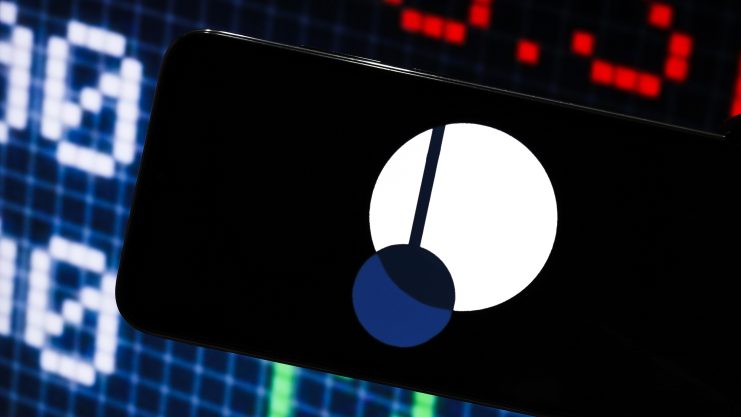 Ein Smartphone mit automatisch gespeichertem Entwurf zeigt das Logo der Kryptowährung Polkadot vor einem digitalen Börsenticker-Bildschirm.