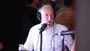 Ein Mann mit grauen Haaren und Bart trägt Kopfhörer und spricht in ein Mikrofon über Bitcoin bei einer Indoor-Veranstaltung.