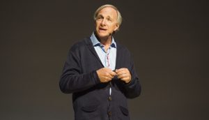 Ein Mann, der ein blaues Hemd und eine dunkle Strickjacke trägt und Ray Dalio ähnelt, spricht auf der Bühne vor einem schlichten, dunklen Hintergrund und hält einen Gegenstand in den Händen, während er seine Prognose für wirtschaftliche Trends erläutert.