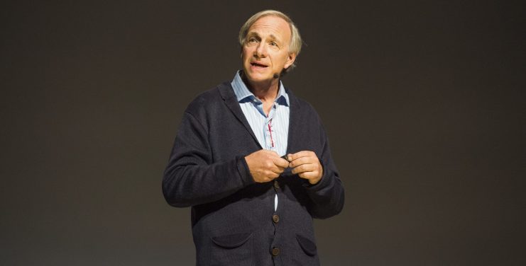 Ein Mann, der ein blaues Hemd und eine dunkle Strickjacke trägt und Ray Dalio ähnelt, spricht auf der Bühne vor einem schlichten, dunklen Hintergrund und hält einen Gegenstand in den Händen, während er seine Prognose für wirtschaftliche Trends erläutert.