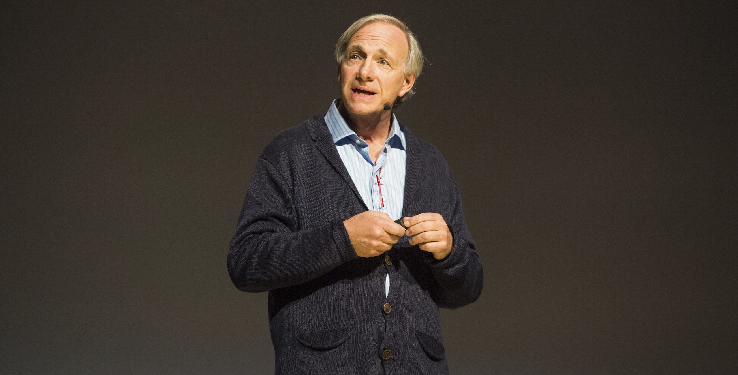 Ein Mann, der ein blaues Hemd und eine dunkle Strickjacke trägt und Ray Dalio ähnelt, spricht auf der Bühne vor einem schlichten, dunklen Hintergrund und hält einen Gegenstand in den Händen, während er seine Prognose für wirtschaftliche Trends erläutert.