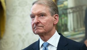 Ein älterer Mann im dunklen Anzug und mit blau gemusterter Krawatte steht in einem Raum und schaut mit neutraler Miene leicht zur Seite, während im Hintergrund Diskussionen über Ripple Spot ETFs widerhallen.