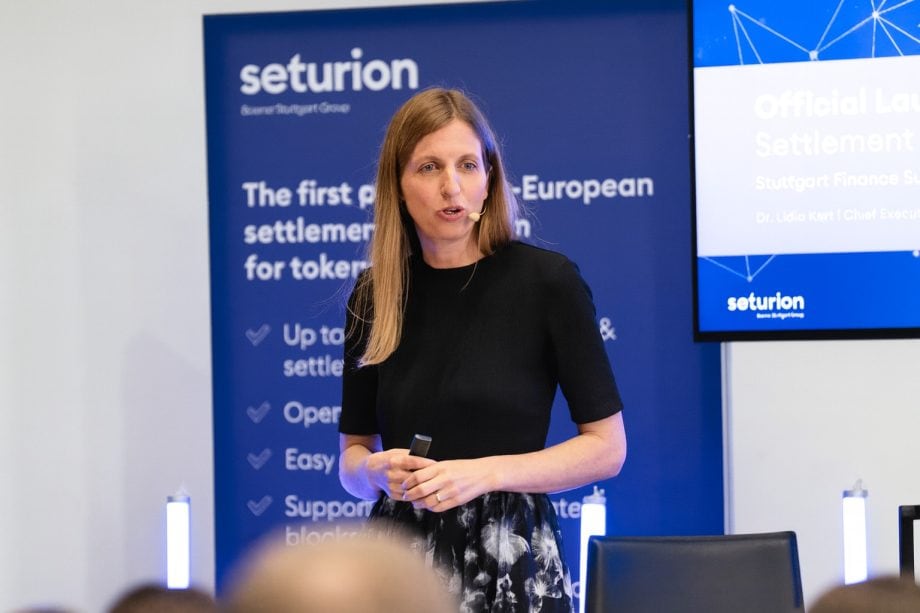 Eine Frau, die ein Mikrofon-Headset trägt, steht und spricht auf einer Veranstaltung, während im Hintergrund ein blaues Seturion-Banner und ein Präsentationsbildschirm mit dem Seturion-Branding zu sehen sind.