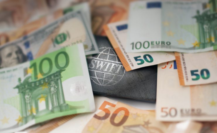 Mehrere Euro- und Dollarscheine umgeben ein metallisches SWIFT-Logo, das internationale Finanztransaktionen, Bankensysteme (Banken) und den wachsenden Einfluss von Stablecoins im globalen Finanzwesen symbolisiert.