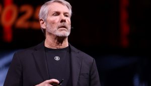 Ein Mann mit grauen Haaren und Bart, der einen schwarzen Blazer und ein schwarzes Hemd mit einem Bitcoin-Logo trägt, spricht auf der Bühne über seine Strategie für den BTC-Kurs und hält ein kleines Gerät in der Hand.