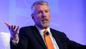 Ein Mann im dunklen Anzug und mit orangefarbener Krawatte gestikuliert mit einer Hand, während er auf einer Veranstaltung über Bitcoin spricht, sitzend vor einem blauen Hintergrund.