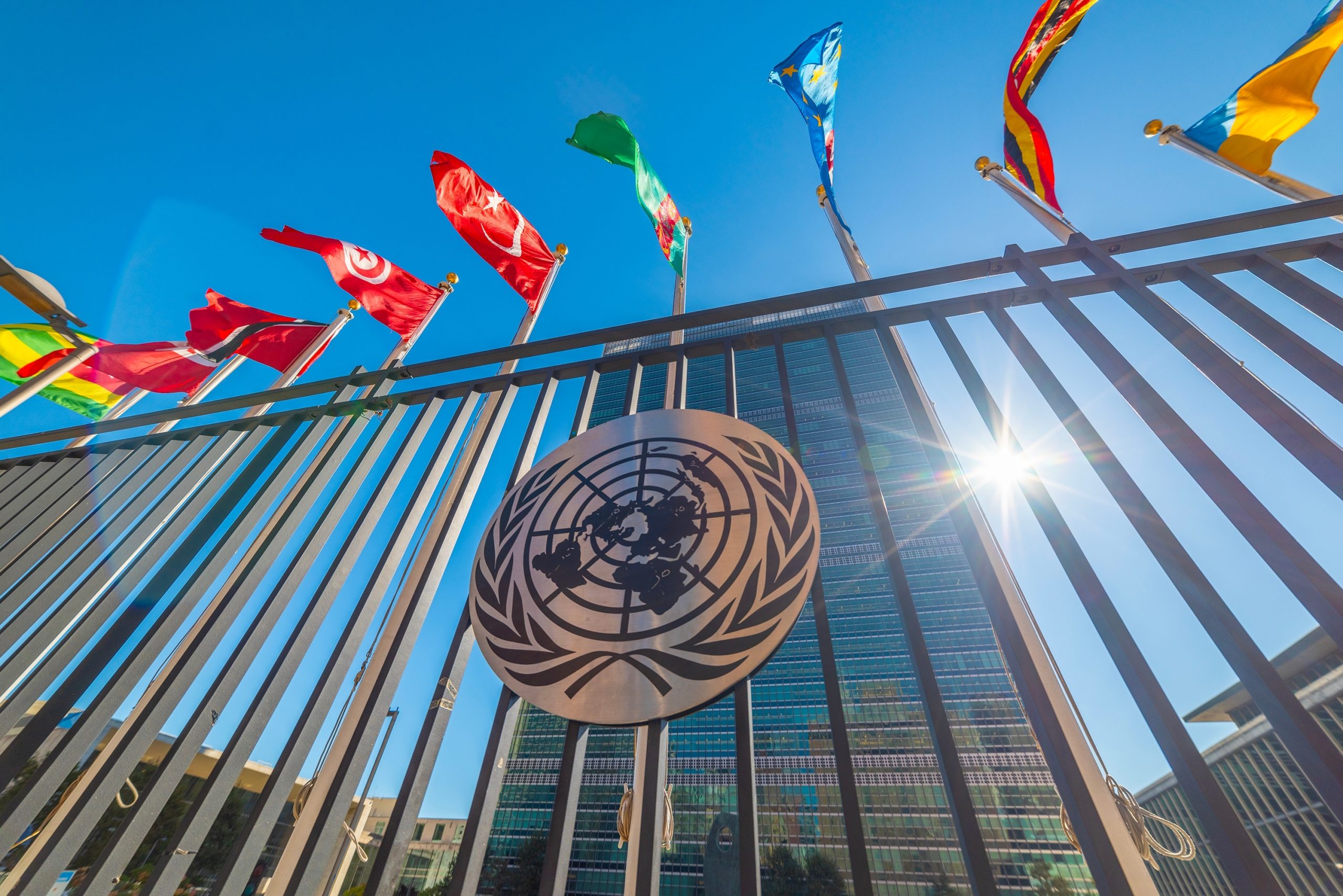 Das Emblem der Vereinten Nationen auf einem Zaun mit mehreren internationalen Flaggen und dem Gebäude des UN-Hauptquartiers unter einem klaren blauen Himmel.