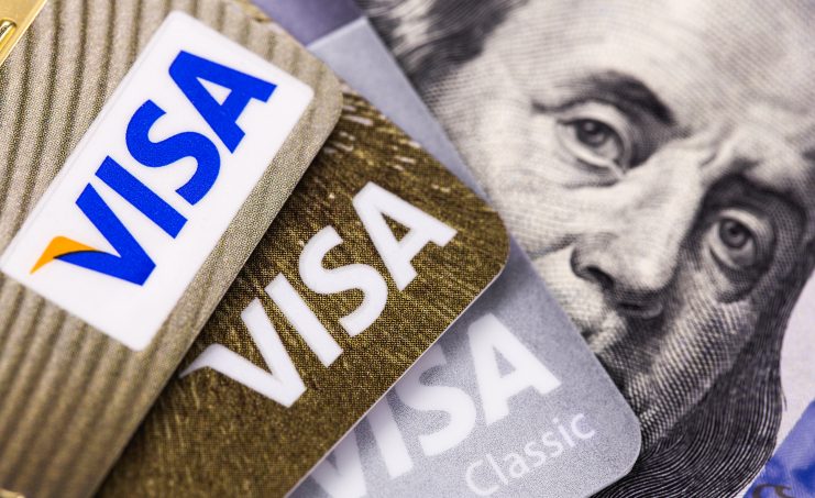 Drei Visa-Kreditkarten sind auf einem amerikanischen Hundert-Dollar-Schein gestapelt, wobei das unverwechselbare Visa-Logo über dem Teilporträt von Benjamin Franklin zu sehen ist.