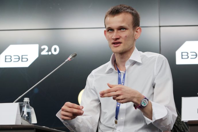 Vitalik Buterin in einem weißen Hemd spricht auf einer Ethereum-Konferenz und gestikuliert mit den Händen. Er sitzt an einem Tisch mit einem Mikrofon und einem Namensschild vor einem Hintergrund mit der Aufschrift 2.0.