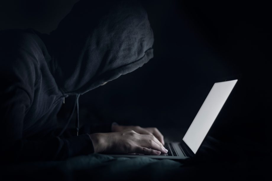 Eine Person, die einen dunklen Kapuzenpulli trägt, benutzt einen Laptop in einer schwach beleuchteten Umgebung, wobei der Bildschirm ihre Hände und ihr Gesicht beleuchtet, während sie sich in Krypto-Transaktionen vertieft.