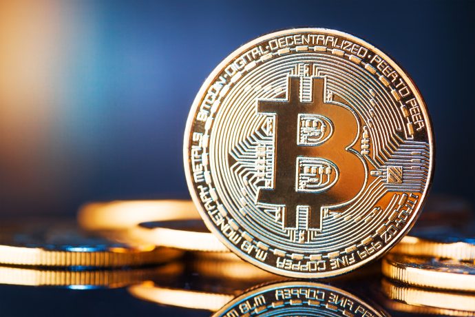 Nahaufnahme eines physischen Bitcoins mit dem Bitcoin-Logo auf einer spiegelnden Oberfläche, mit anderen Münzen im Hintergrund, die leicht verschwommen sind.