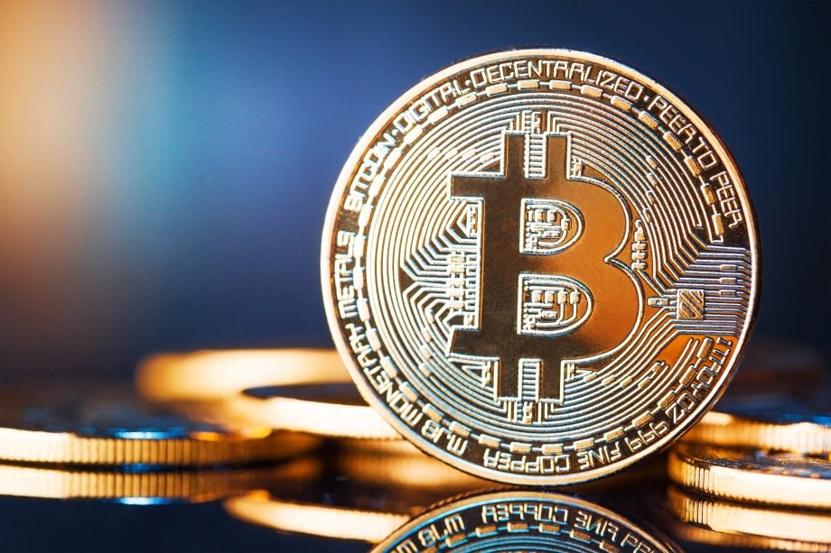 Nahaufnahme eines physischen Bitcoins mit dem Bitcoin-Logo auf einer spiegelnden Oberfläche, mit anderen Münzen im Hintergrund, die leicht verschwommen sind.